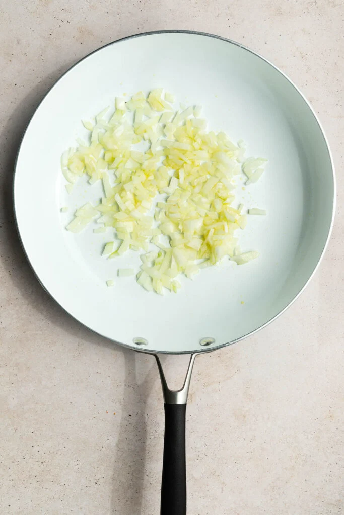 A white frying pan with chopped onions spread in the center, ready to be sautéed as the first step in a creamy vodka sauce recipe, rests on a light beige countertop.