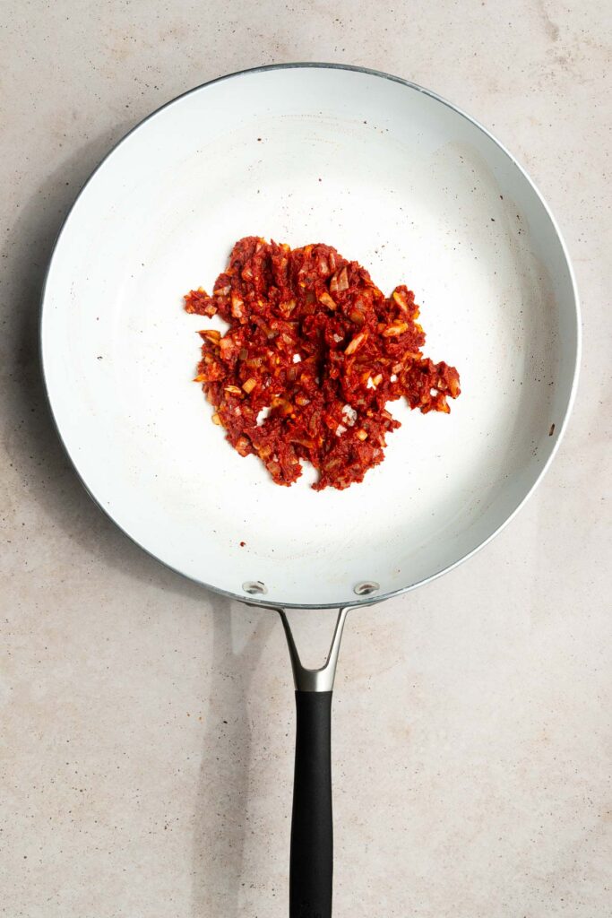 A white frying pan with a black handle contains a small pile of finely chopped, sautéed red ingredients, possibly tomatoes and onions—perfect as the base for a delicious vodka sauce recipe—on a light-colored surface.