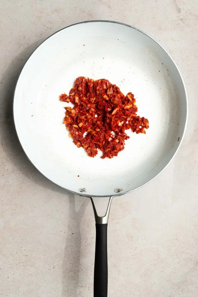 A white frying pan with a black handle contains a small pile of finely chopped, sautéed red ingredients, possibly tomatoes and onions—perfect as the base for a delicious vodka sauce recipe—on a light-colored surface.