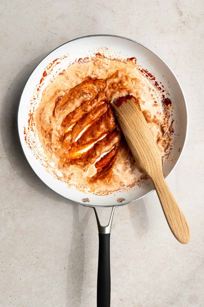 A white frying pan with a black handle contains a creamy, red-orange vodka sauce being stirred with a wooden spatula on a light countertop. The partially mixed and slightly thick sauce is perfect for trying out your favorite vodka sauce recipe.