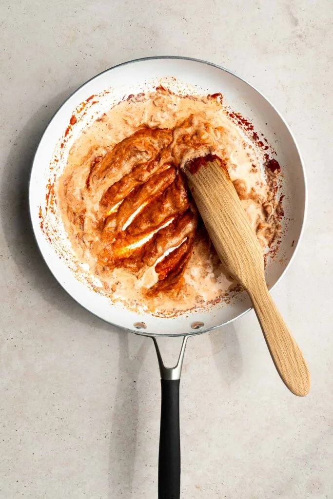 A white frying pan with a black handle contains a creamy, red-orange vodka sauce being stirred with a wooden spatula on a light countertop. The partially mixed and slightly thick sauce is perfect for trying out your favorite vodka sauce recipe.