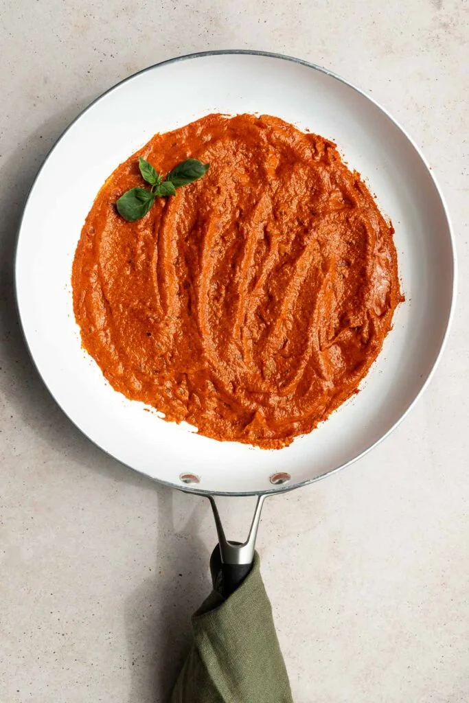 A white skillet filled with creamy red vodka sauce recipe, garnished with a sprig of fresh basil, sits on a light countertop. The handle of the skillet is wrapped with a green cloth.