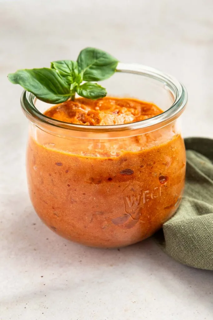 A glass jar filled with creamy copycat Rao's vodka sauce, topped with fresh basil leaves, sits on a light surface next to a folded green cloth.