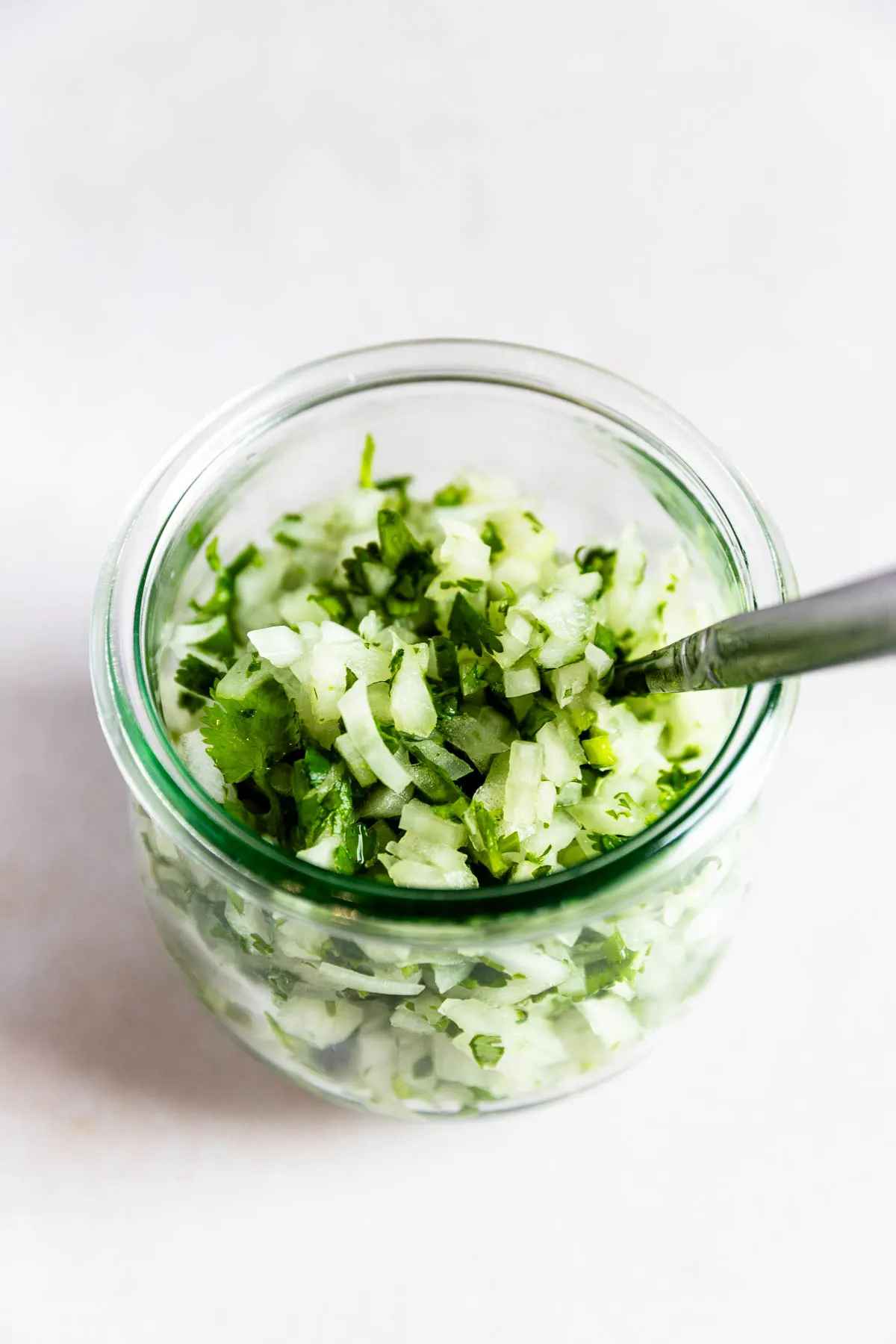 A glass jar filled with finely chopped onions and cilantro, with a spoon inside, placed on a light-colored surface.