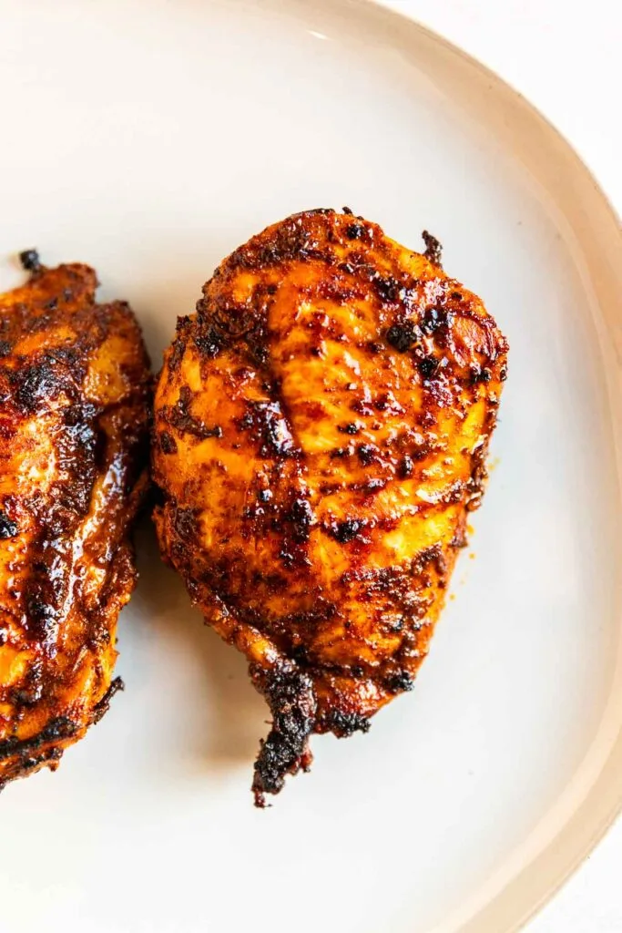 A close-up of two pieces of grilled chicken breast with a golden brown, slightly charred exterior, served on a light-colored plate for ancho chile chicken.