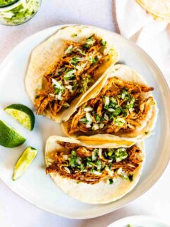 Three soft corn tortillas filled with shredded ancho chicken tacos, topped with chopped onions and cilantro, are arranged on a white plate with lime wedges on the side.