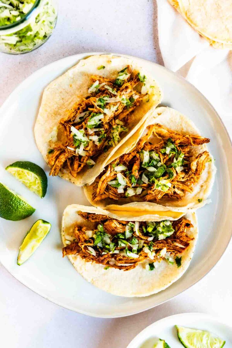 Three soft corn tortillas filled with shredded ancho chicken tacos, topped with chopped onions and cilantro, are arranged on a white plate with lime wedges on the side.