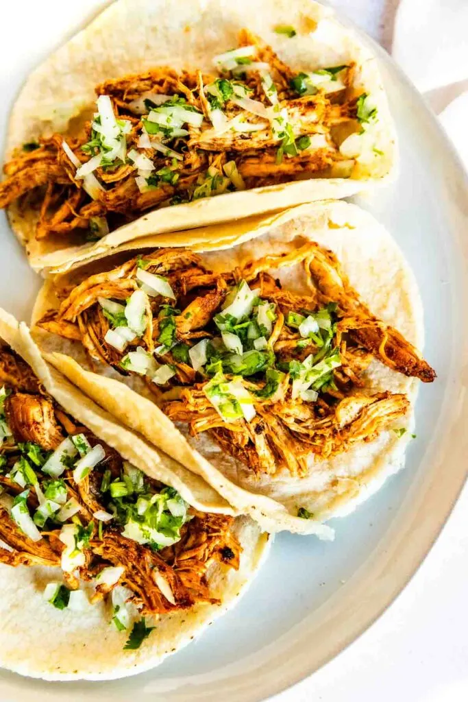Three ancho chicken tacos filled with shredded seasoned meat, topped with chopped onions and fresh cilantro, served on soft corn tortillas on a white plate.