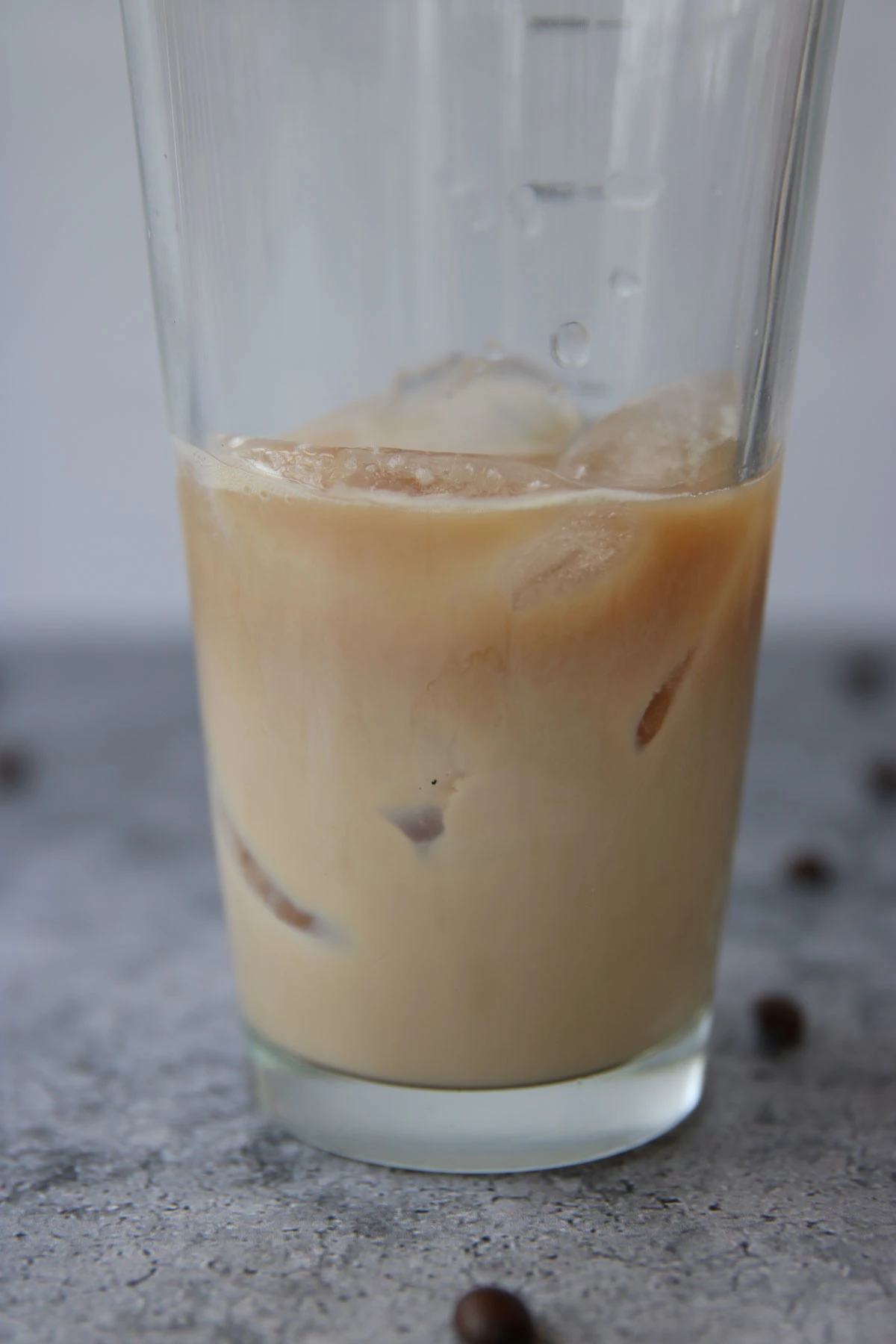 A close-up of a glass filled with iced coffee, showing ice cubes and creamy coffee swirled together, evokes the look of a Baileys espresso martini, placed on a gray surface with scattered coffee beans in the background.