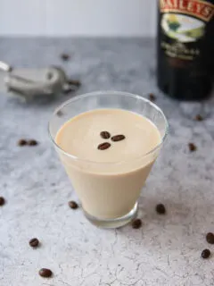 A glass of creamy coffee liqueur, inspired by an espresso martini recipe with Baileys, is topped with three coffee beans on a gray textured surface. Scattered beans and a blurred Baileys bottle and cocktail strainer appear in the background.