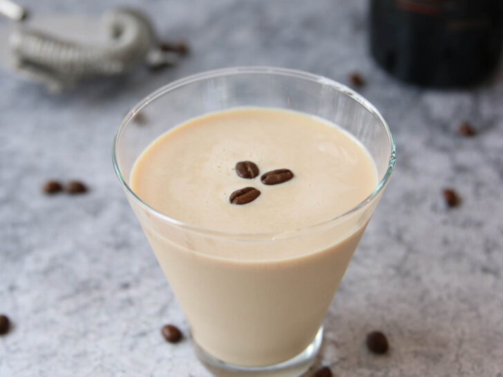 A glass of creamy coffee liqueur, inspired by an espresso martini recipe with Baileys, is topped with three coffee beans on a gray textured surface. Scattered beans and a blurred Baileys bottle and cocktail strainer appear in the background.