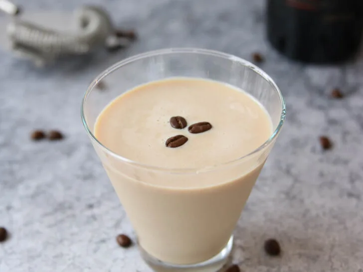 A glass of creamy coffee liqueur, inspired by an espresso martini recipe with Baileys, is topped with three coffee beans on a gray textured surface. Scattered beans and a blurred Baileys bottle and cocktail strainer appear in the background.