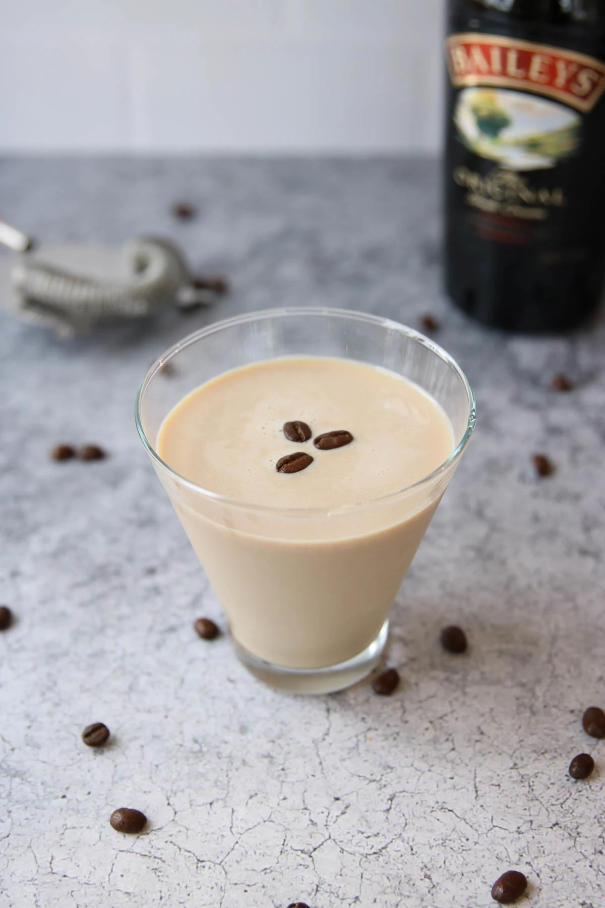 A glass of creamy coffee liqueur, inspired by an espresso martini recipe with Baileys, is topped with three coffee beans on a gray textured surface. Scattered beans and a blurred Baileys bottle and cocktail strainer appear in the background.