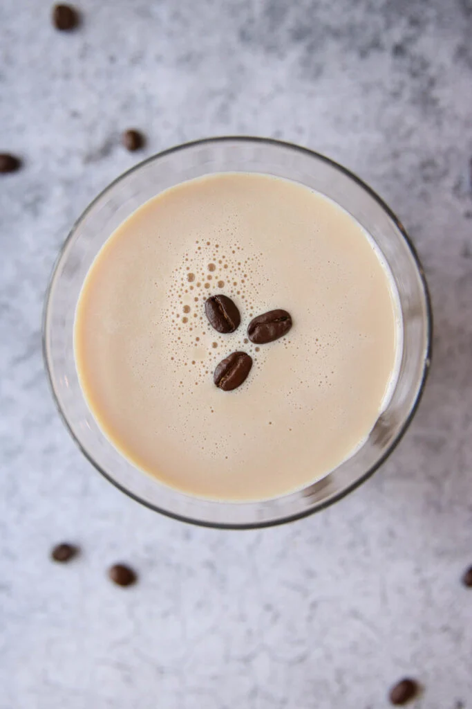 A glass of creamy, coffee-colored espresso martini with Baileys, topped with three coffee beans, viewed from above, with a few coffee beans scattered on a light, textured surface in the background.