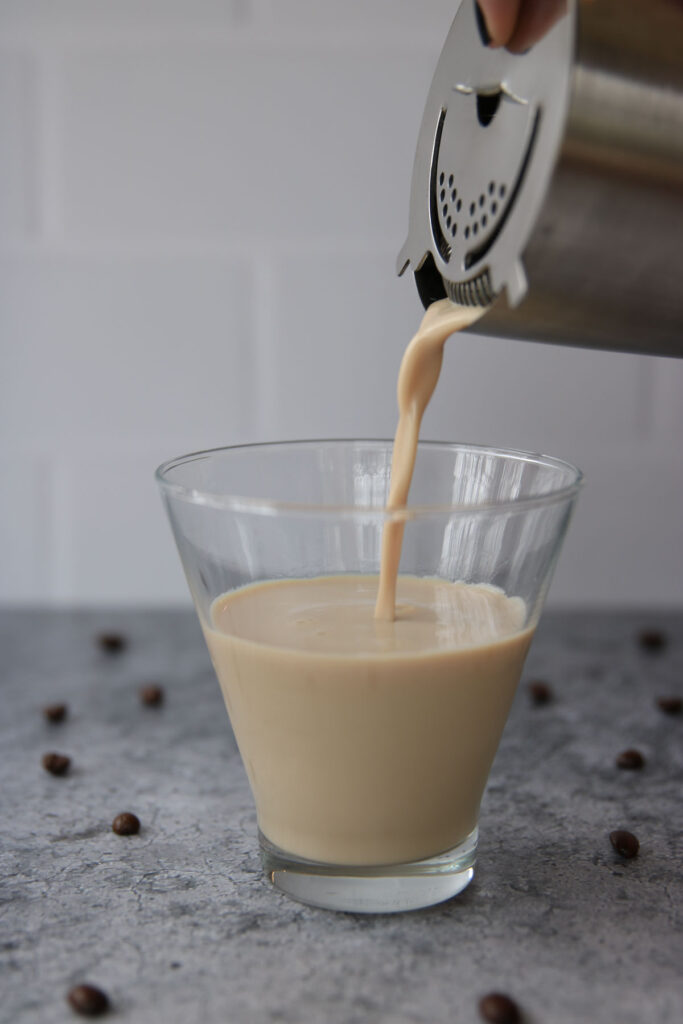 A creamy beige Baileys espresso martini is being poured from a cocktail shaker into a clear glass. Coffee beans are scattered on a gray surface, and the background is a white tiled wall.