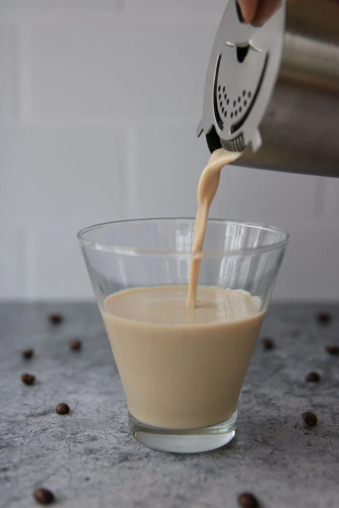 A creamy beige Baileys espresso martini is being poured from a cocktail shaker into a clear glass. Coffee beans are scattered on a gray surface, and the background is a white tiled wall.