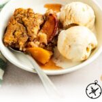 A white bowl filled with a serving of peach cobbler and two scoops of vanilla ice cream, with a spoon resting inside. The cobbler has a golden-brown crust and tender peach slices. Text below reads: “Easy Peach Cobbler.”.