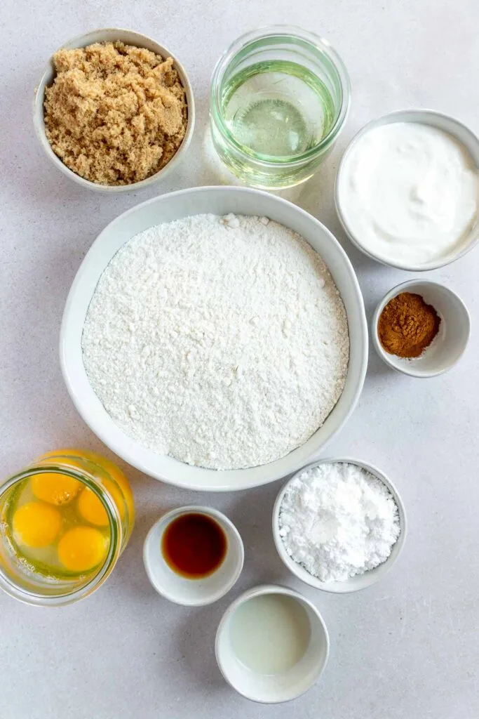 A top view of baking ingredients in bowls: flour, brown sugar, vegetable oil, yogurt, ground cinnamon, eggs, vanilla extract, powdered sugar, and a small amount of liquid on a white surface.