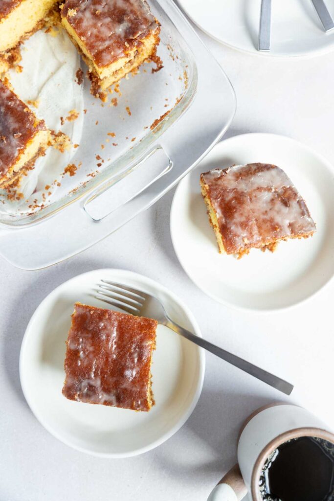 Glazed square pieces of honey bun cake on white plates, with a glass baking dish containing more cake in the background, and a cup of coffee nearby on a white surface.