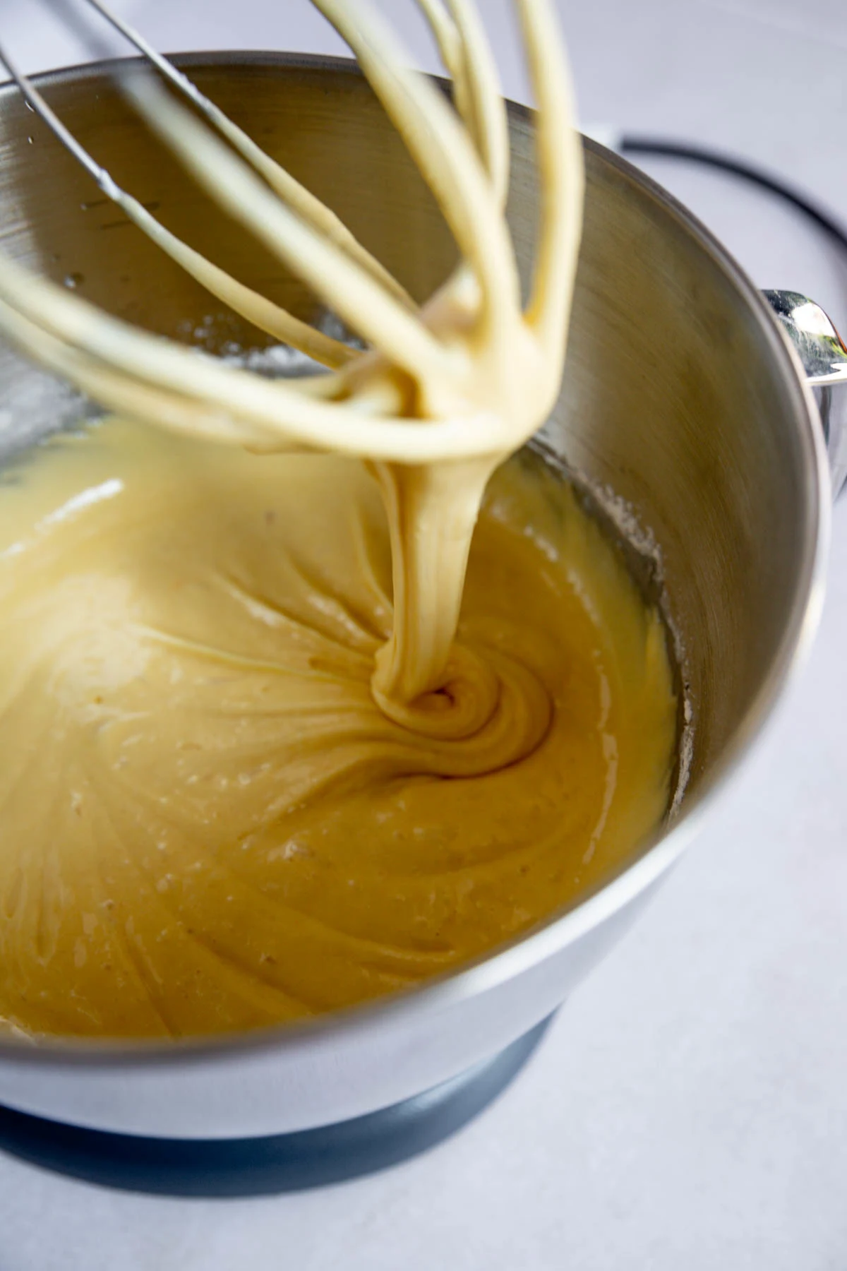 A close-up of thick yellow batter being mixed in a stainless steel bowl with a metal whisk attachment, showing a smooth, creamy texture.