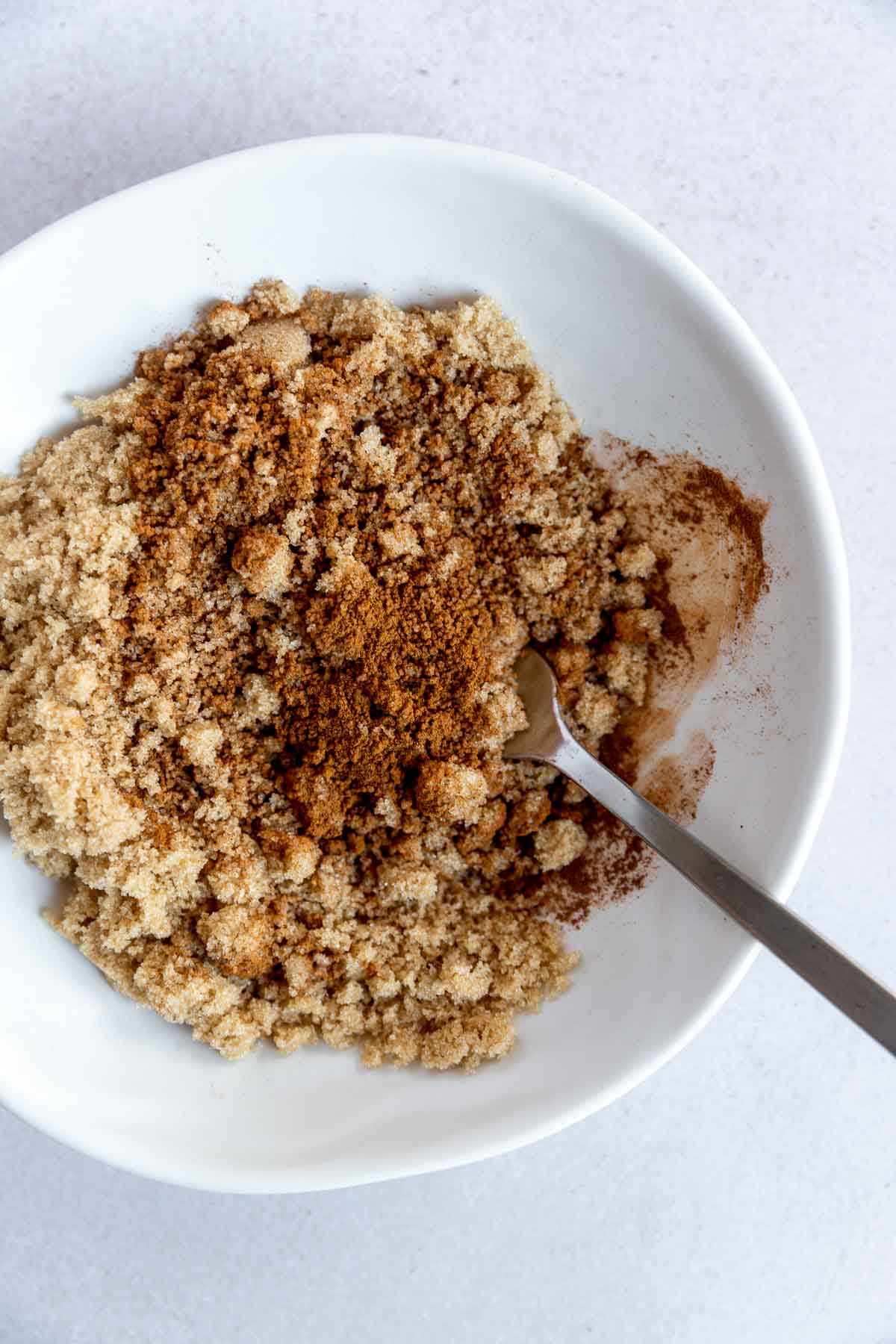 A white bowl filled with light brown sugar and a sprinkle of ground cinnamon, with a metal fork resting inside on a white surface.