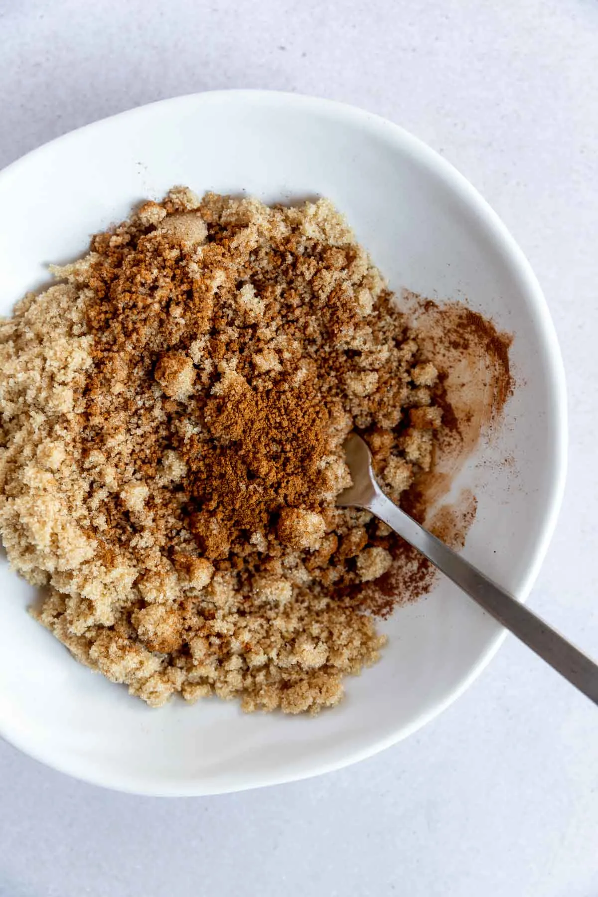 A white bowl filled with light brown sugar and a sprinkle of ground cinnamon, with a metal fork resting inside on a white surface.