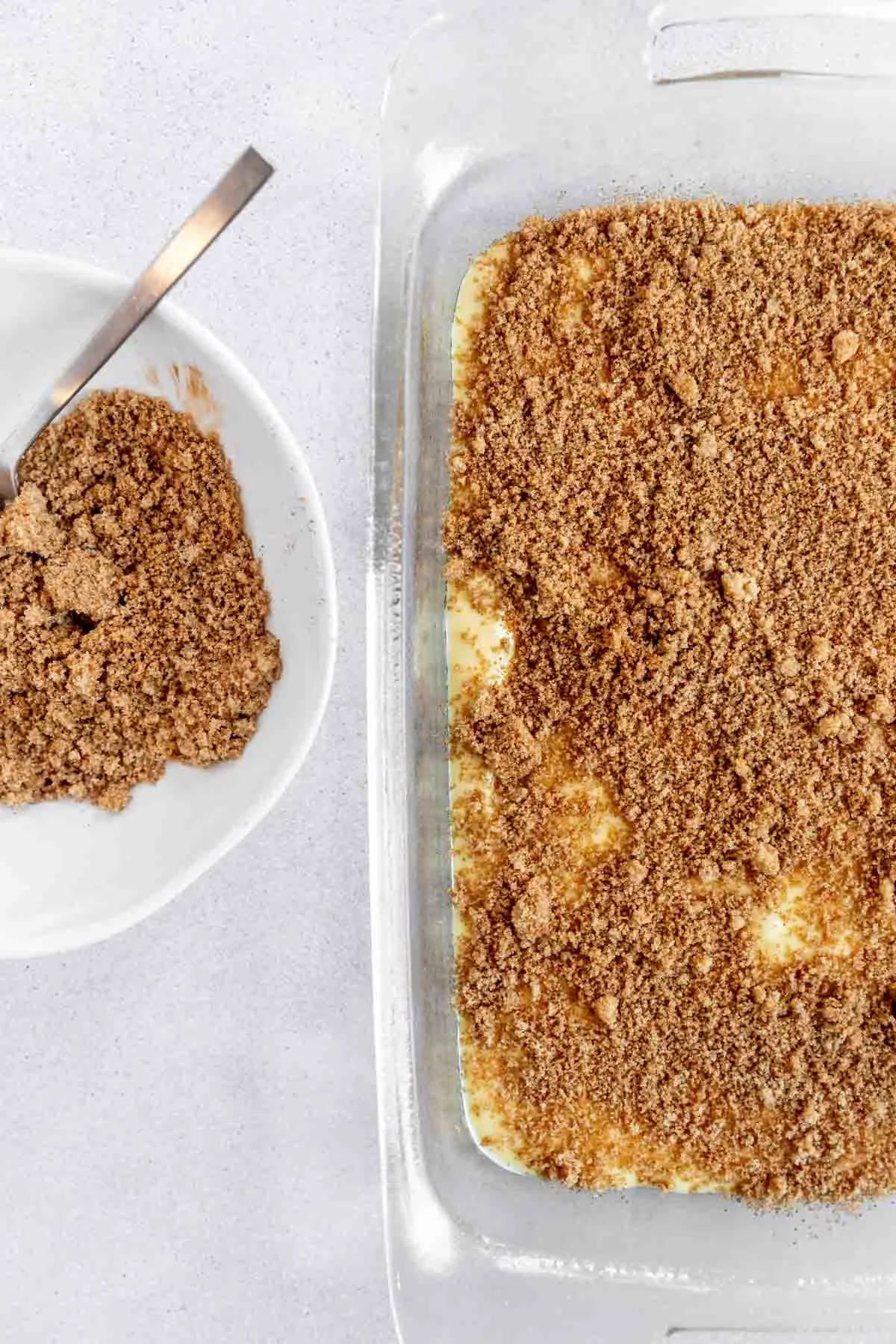 A glass baking dish filled with an unbaked dessert topped with a crumbly brown streusel. Next to it, a white bowl holds extra streusel mixture with a spoon inside. The background is a light-colored surface.