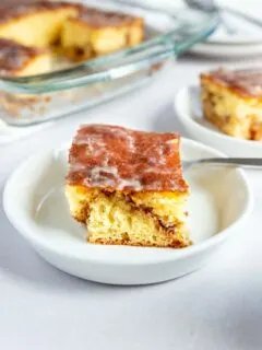 A square slice of glazed honey bun cake sits on a white plate with a fork, while the rest of the cinnamon roll cake waits in a glass baking dish in the background.