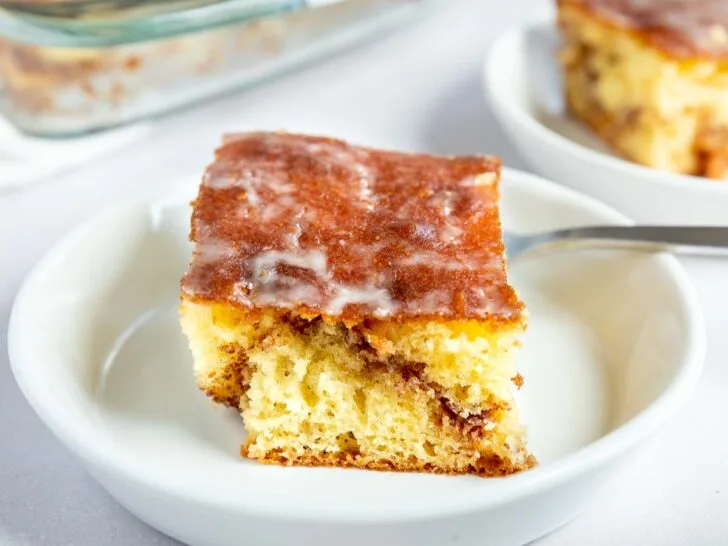 A square slice of glazed honey bun cake sits on a white plate with a fork, while the rest of the cinnamon roll cake waits in a glass baking dish in the background.