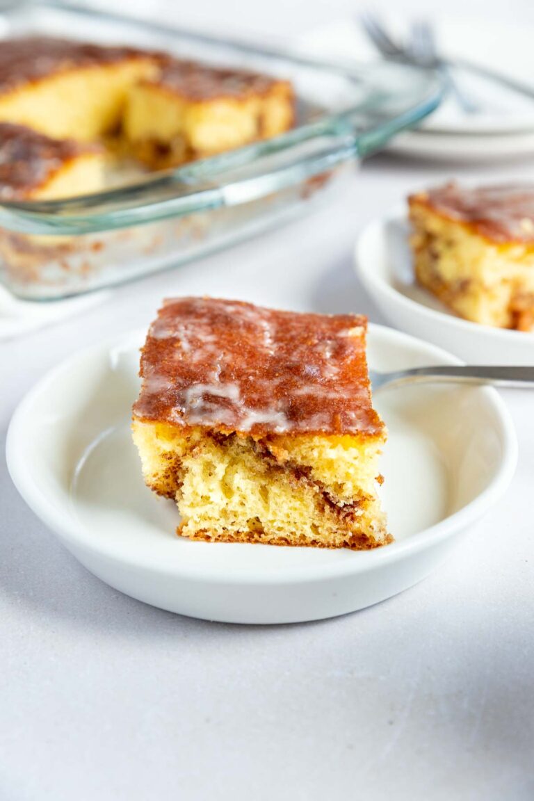 A square slice of glazed honey bun cake sits on a white plate with a fork, while the rest of the cinnamon roll cake waits in a glass baking dish in the background.