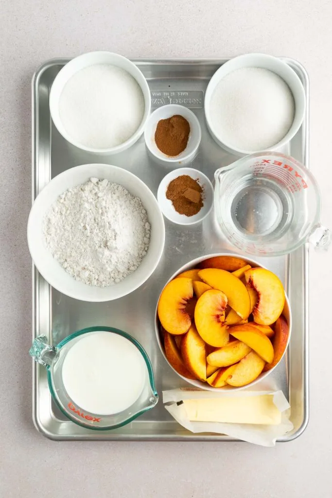 A metal tray holds bowls of flour, sugar, cinnamon, and sliced peaches, along with a measuring cup of milk, a measuring cup of water, and two sticks of butter, arranged neatly on a light countertop.