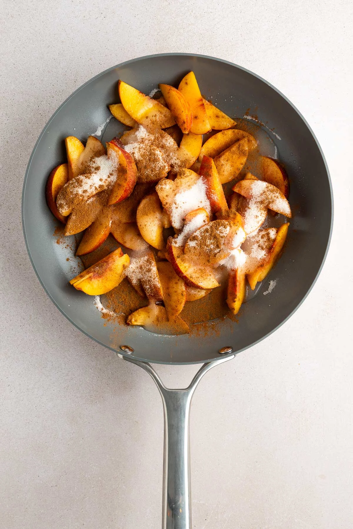 A frying pan filled with sliced apples topped with cinnamon, sugar, and chunks of butter, ready to be cooked on a light-colored countertop.