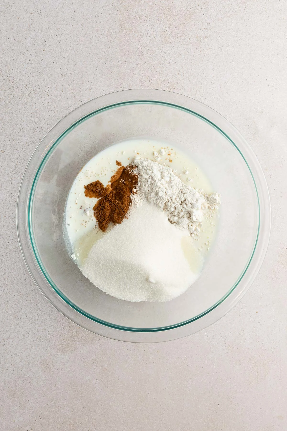A glass mixing bowl on a light surface containing milk, flour, white sugar, and ground cinnamon, with the ingredients separated and not yet mixed together.