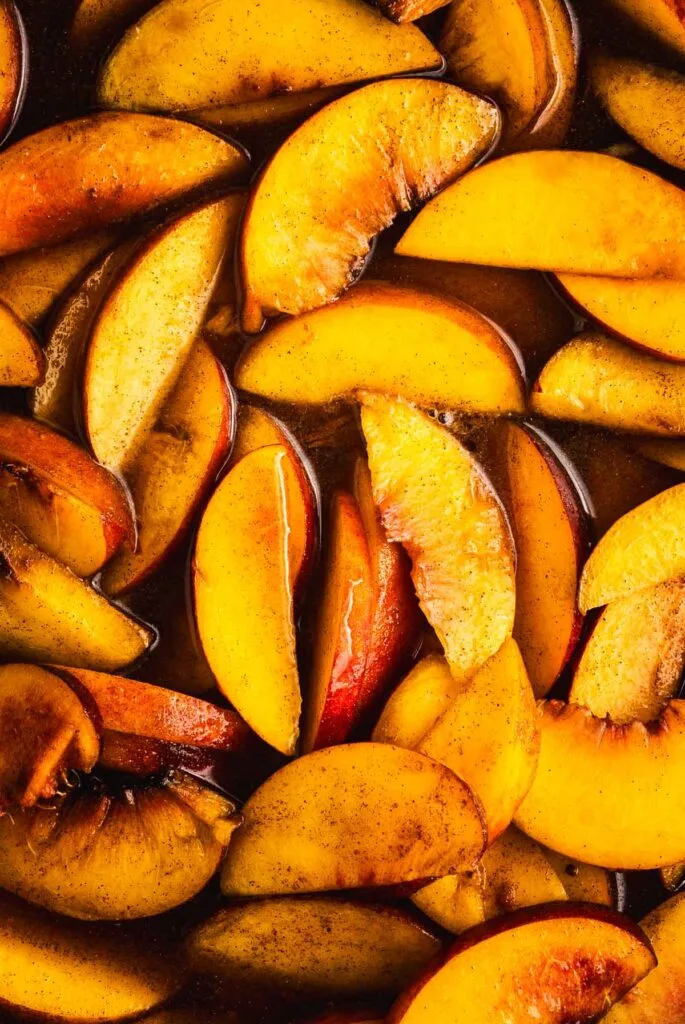 Sliced fresh peaches coated with cinnamon and sugar, close-up. The yellow-orange fruit pieces are arranged closely together, some with red skin visible, and appear juicy and glossy.