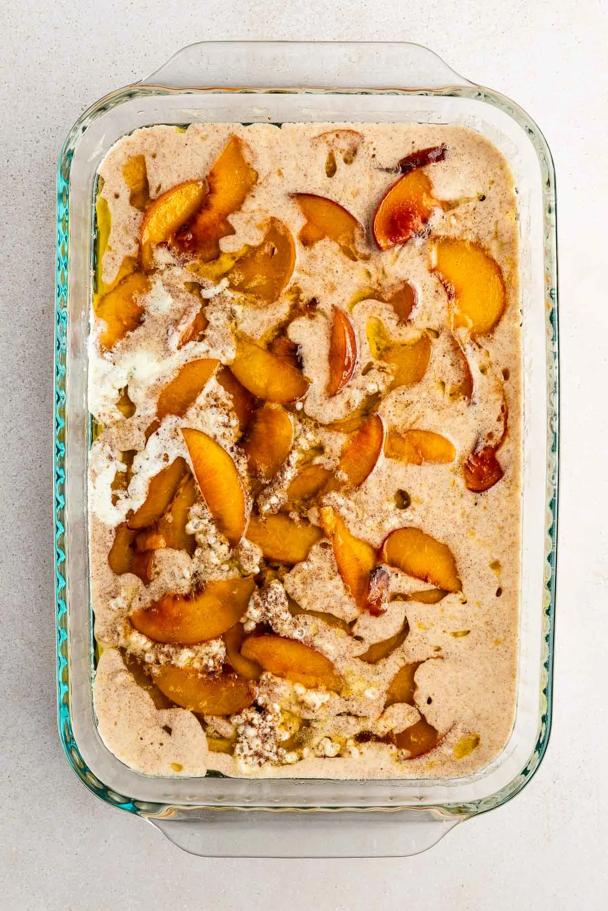 A glass baking dish filled with sliced peaches mixed into a creamy, cinnamon-speckled batter, ready to be baked into a peach cobbler.