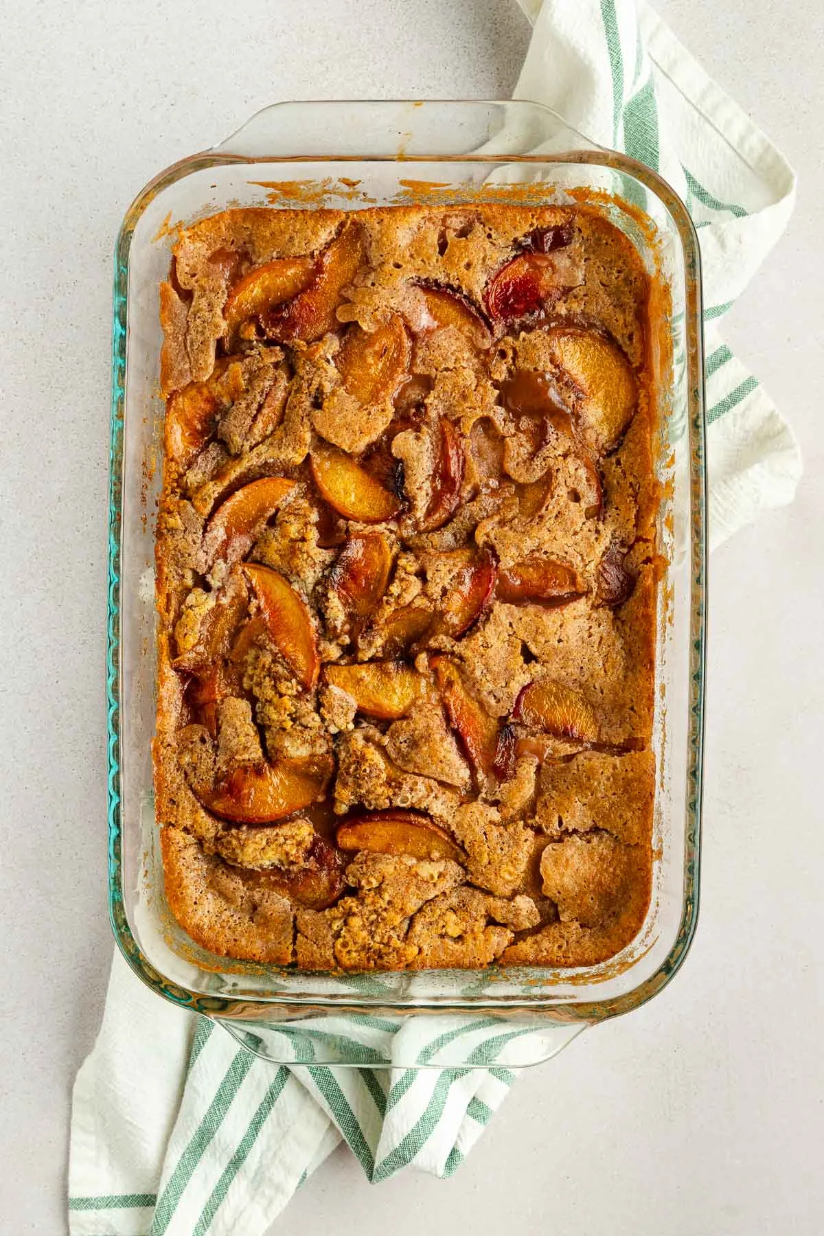 A glass baking dish filled with golden-brown peach cobbler sits on a white surface, with a green and white striped towel partially underneath. Juicy slices of peaches and baked crust are visible on top of the peach cobbler.