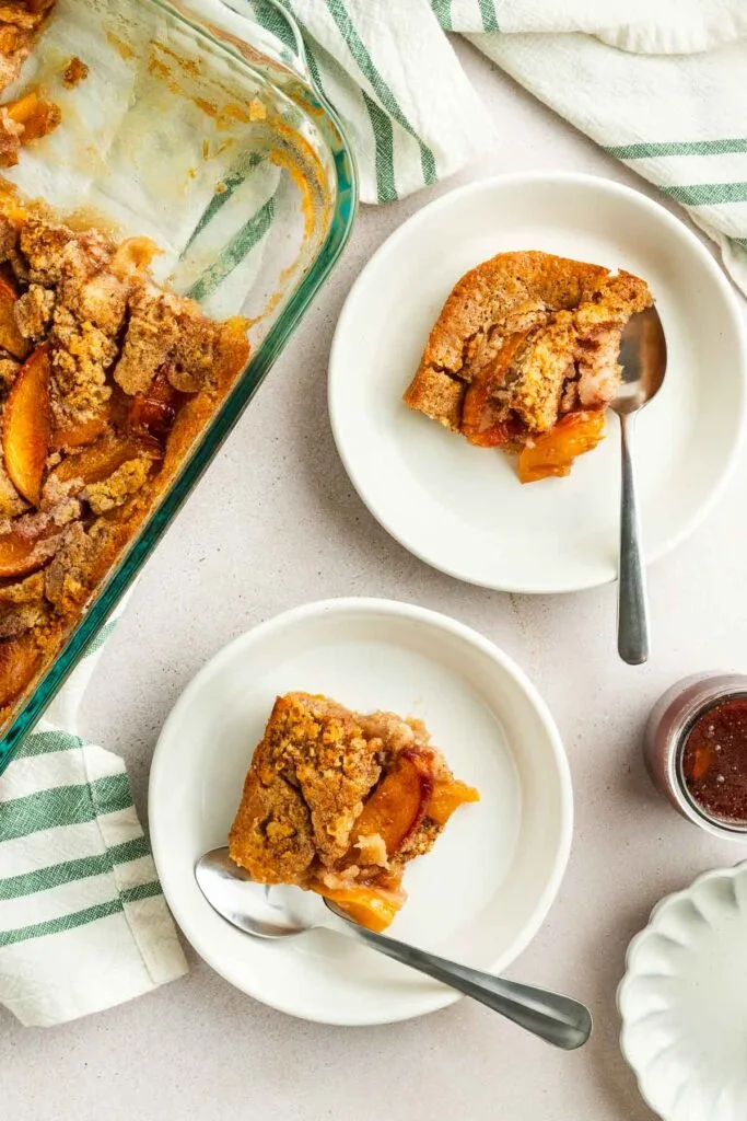 Overhead view of two plates with peach cobbler and spoons, next to a baking dish filled with more peach cobbler. A striped towel and a small jar of jam rest nearby on a light-colored surface.