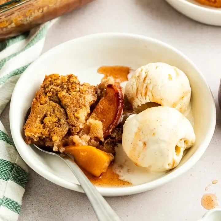 A white bowl with a serving of fruit cobbler and two scoops of vanilla ice cream, made from an easy peach cobbler recipe, with a spoon resting inside. A baking dish and another dessert plate appear in the background.