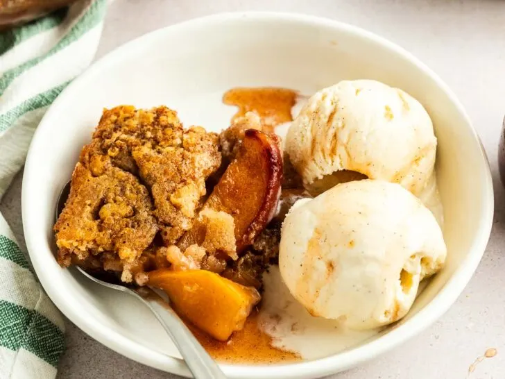 A white bowl with a serving of fruit cobbler and two scoops of vanilla ice cream, made from an easy peach cobbler recipe, with a spoon resting inside. A baking dish and another dessert plate appear in the background.