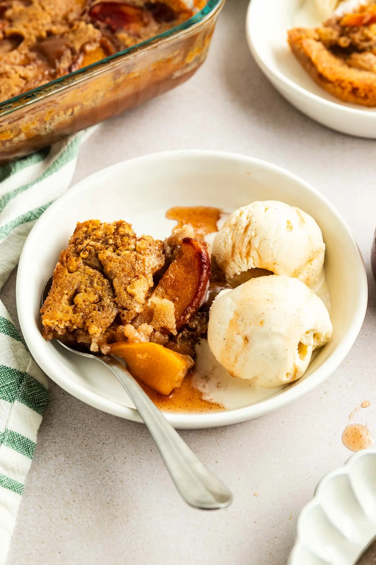 A white bowl with a serving of fruit cobbler and two scoops of vanilla ice cream, made from an easy peach cobbler recipe, with a spoon resting inside. A baking dish and another dessert plate appear in the background.