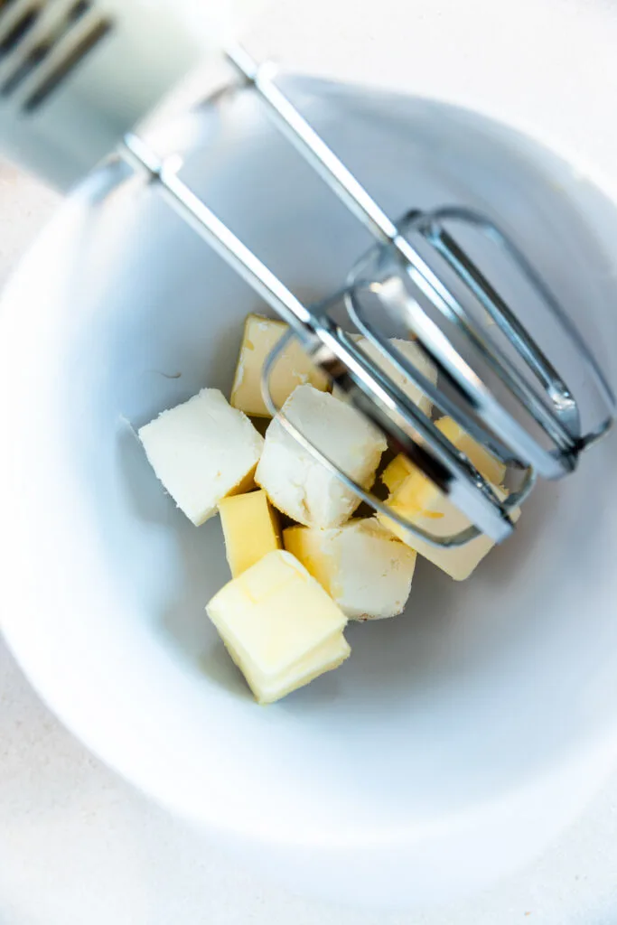 Several cubes of butter and cream cheese in a white mixing bowl with metal beaters from a hand mixer positioned above them, ready for mixing.