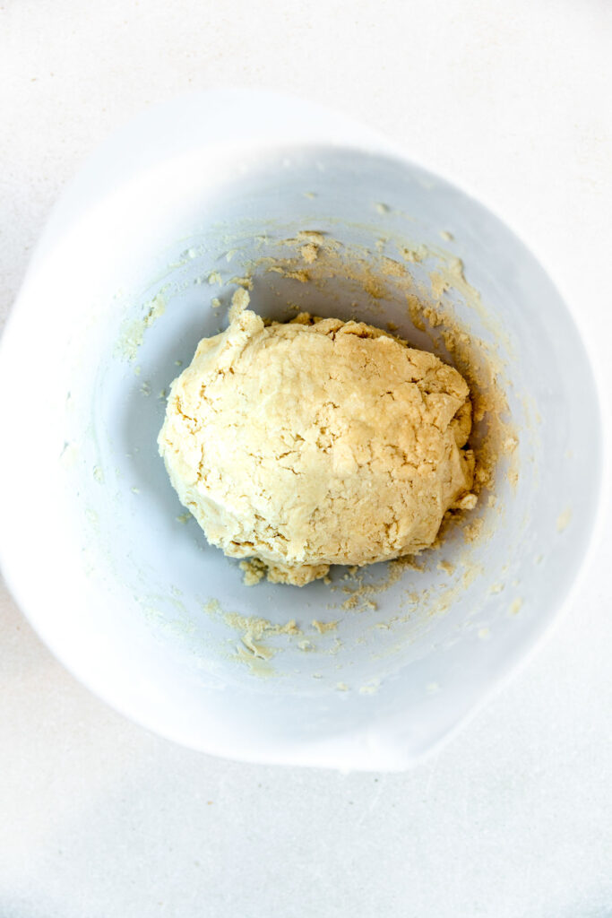 A ball of pale, yellowish dough sits in the center of a white mixing bowl on a light countertop. Some dough residue is visible on the sides of the bowl.