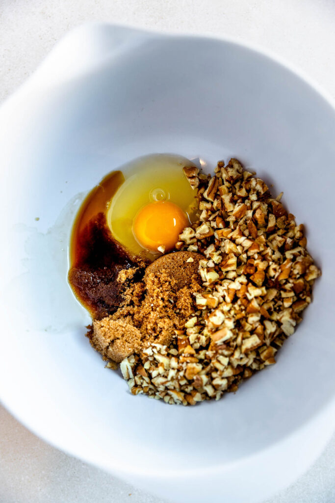 A white mixing bowl containing a raw egg, chopped pecans, brown sugar, and vanilla extract all unmixed and arranged in separate sections.