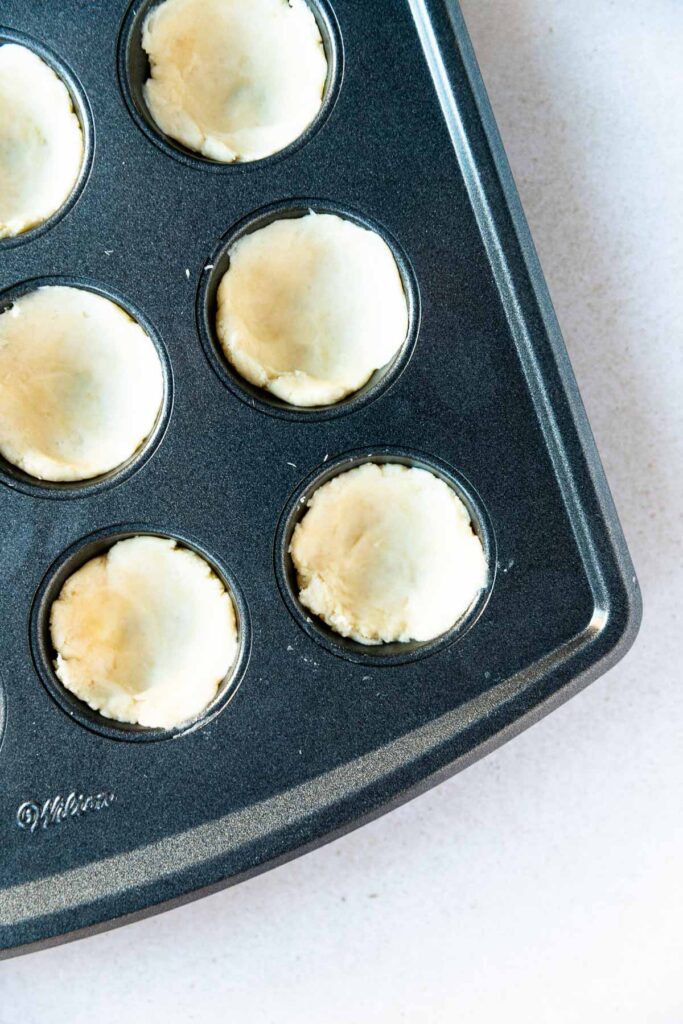 A close-up of a nonstick mini muffin pan filled with rounds of raw pecan tassie dough, pressed into each cup, ready for baking. The pan is on a light-colored surface.