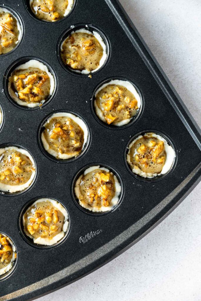 A close-up of a black muffin tin filled with easy pecan tassies. The mini tarts have a creamy crust and nut-filled centers, ready to be baked. The background is a light-colored countertop.