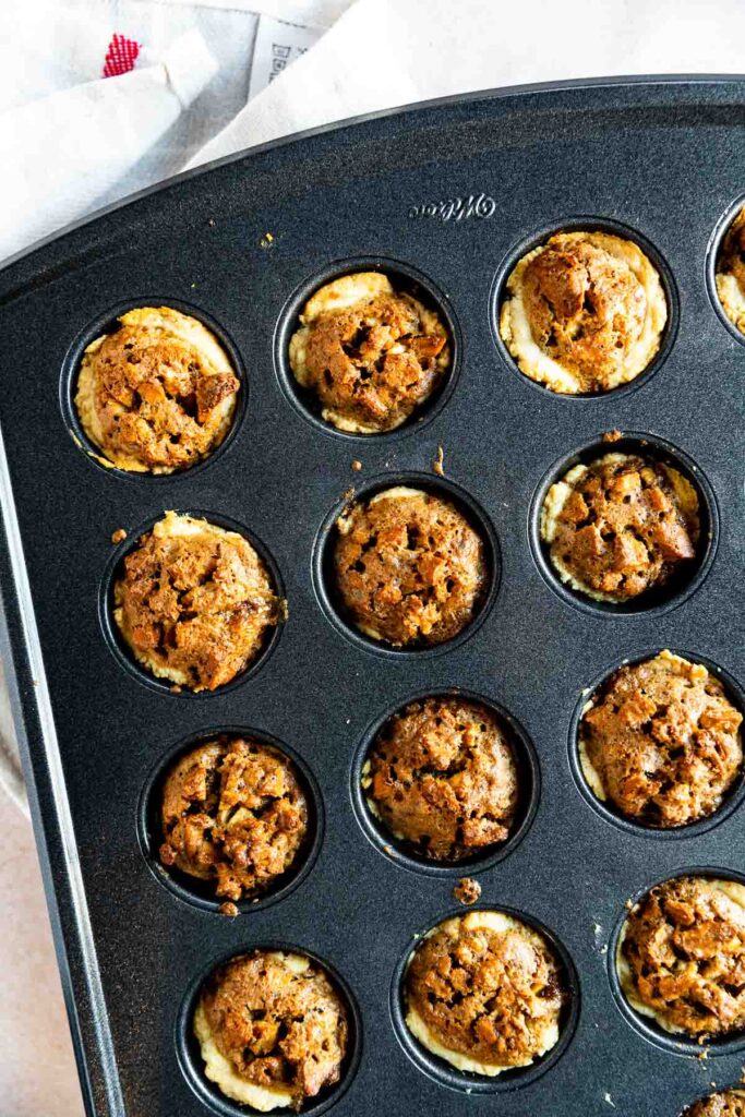 A muffin tin filled with twelve baked mini cheesecakes, each topped with a crumbly brown streusel reminiscent of a pecan tassie recipe, rests on a light surface with part of a white cloth in the background.