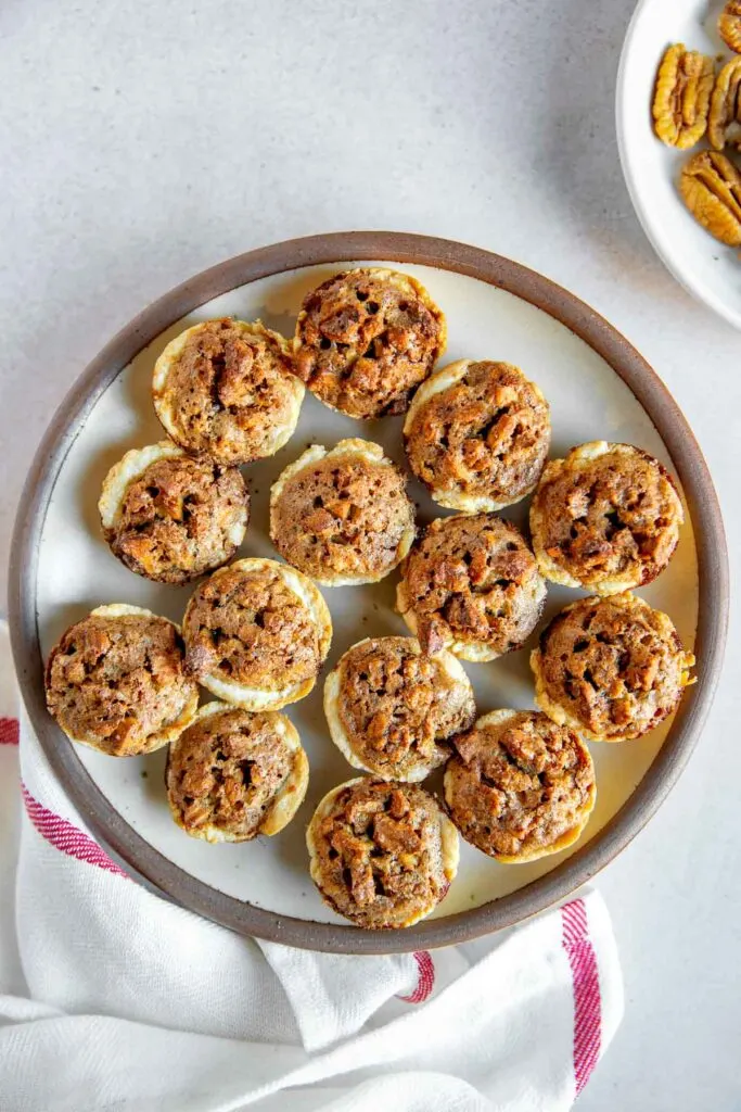 A plate of bite-sized pecan tassies arranged in a circular pattern, viewed from above, with a red-striped cloth underneath and a small dish of pecans in the corner.