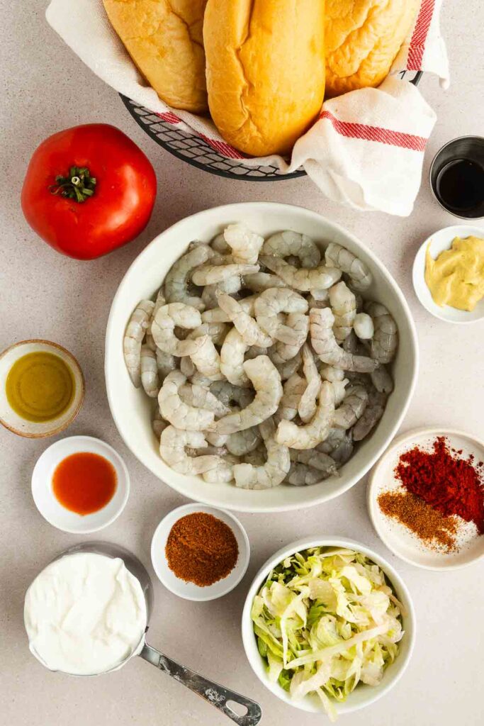 Ingredients for shrimp poboys are arranged on a countertop: a bowl of raw shrimp, sandwich rolls, a tomato, shredded lettuce, seasonings, sauces, and a cup of greek yogurt.