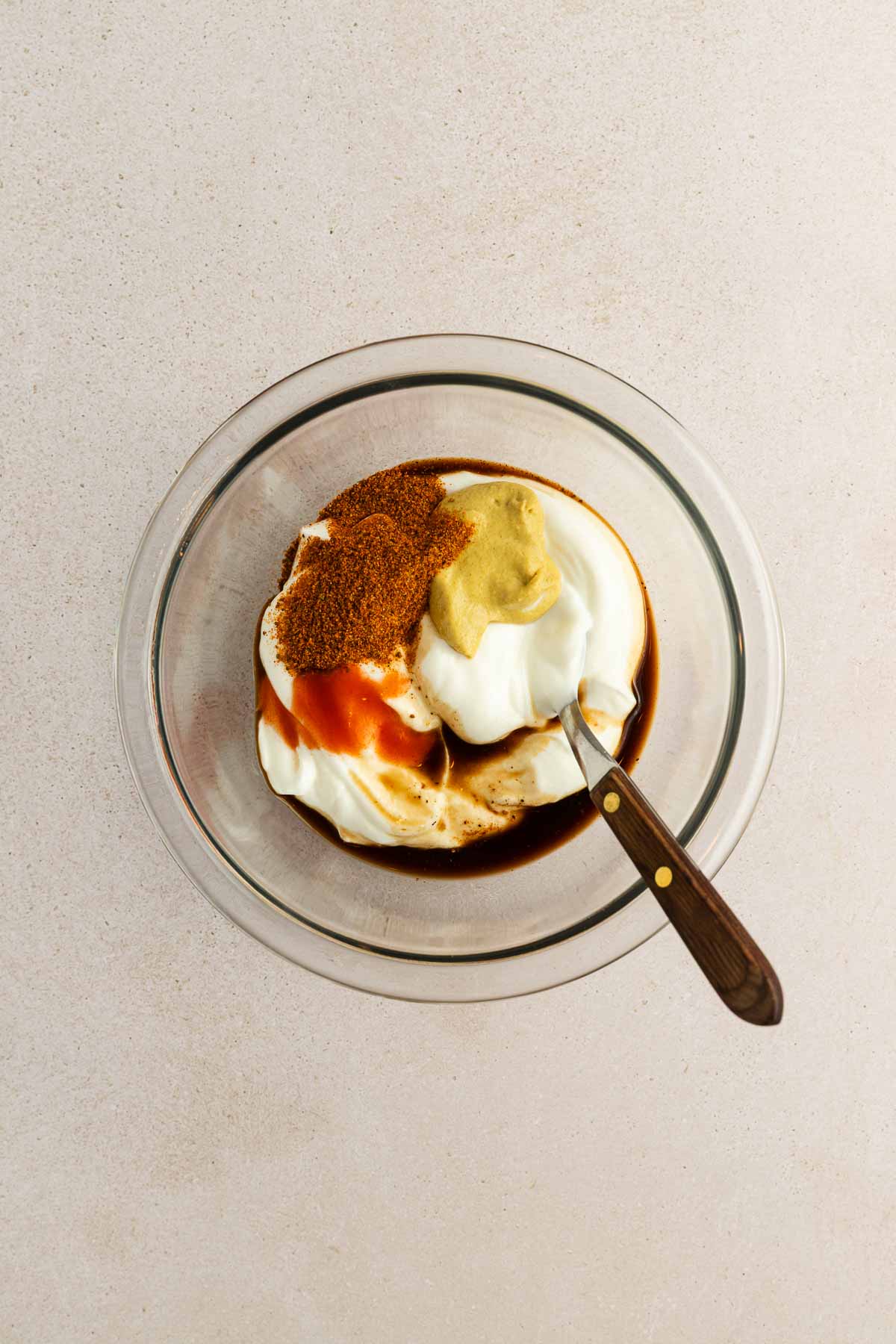 A glass bowl containing yogurt, old bay, mustard, hot sauce, and Worcestershire sauce, with a spoon resting inside, on a light-colored surface.