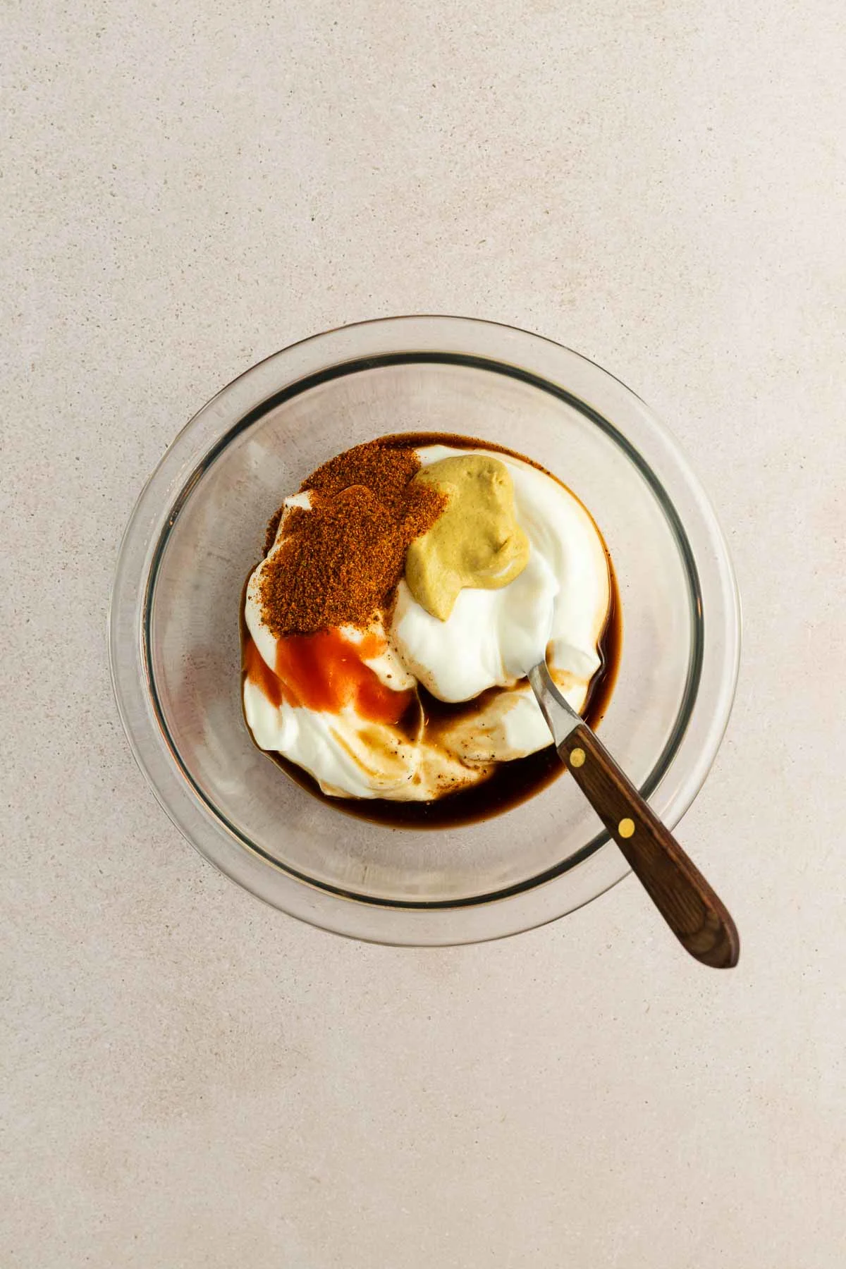 A glass bowl containing yogurt, old bay, mustard, hot sauce, and Worcestershire sauce, with a spoon resting inside, on a light-colored surface.